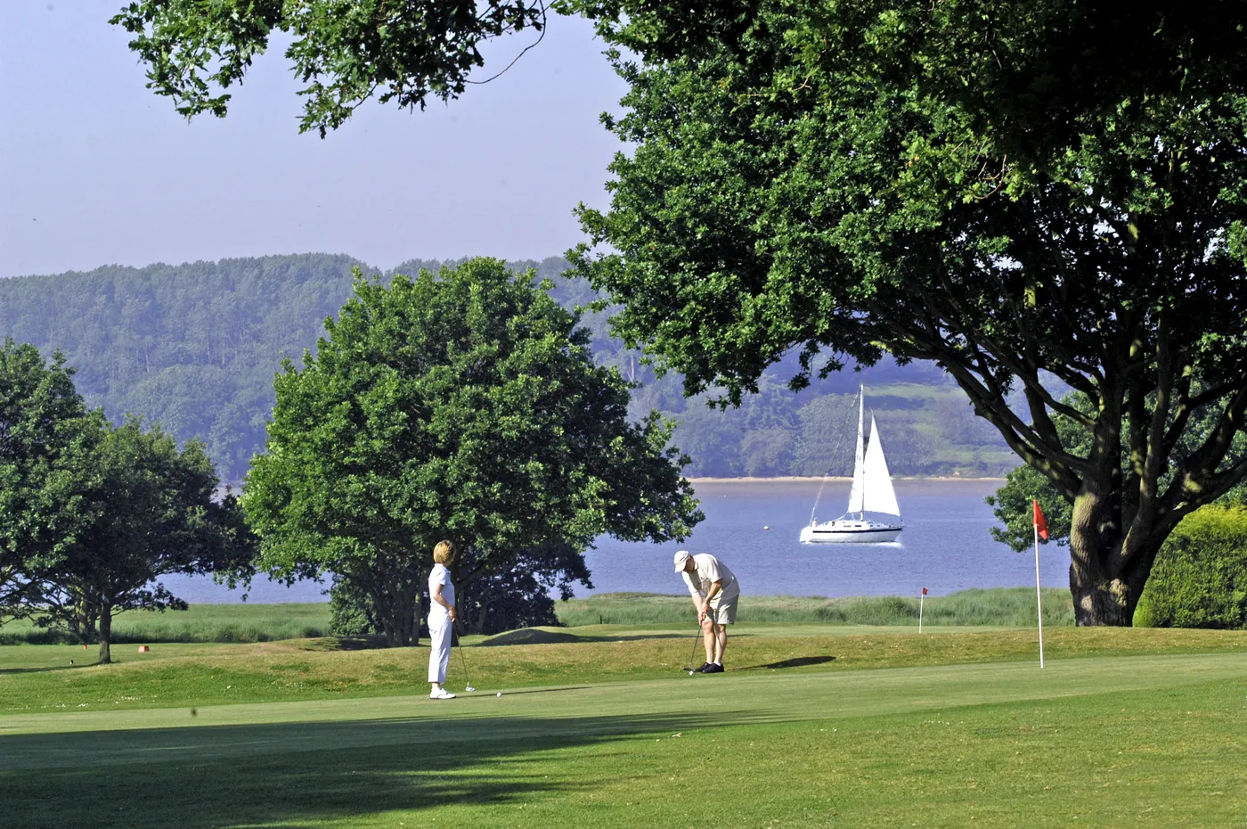 Golfers on the green at Alnesbourne Priory Golf Course with River Orwell and sailing boats in the background, near Ipswich Suffolk